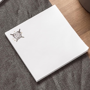 May include: A stack of white square sticky notes with a black and gold geometric design in the upper left corner. The notes are on a dark gray surface, with a laptop and pen visible in the background.