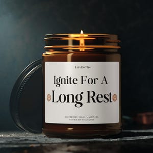 May include: A lit candle in a brown glass jar with a gold lid. The label reads "Ignite For A Long Rest" in a serif font. The candle is on a dark surface with a dark background.