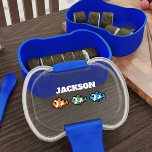 May include: A blue bento box with a clear lid, containing sushi rolls. The lid is printed with the name "JACKSON" and pixelated fish graphics. A blue spoon and another blue container are also visible.