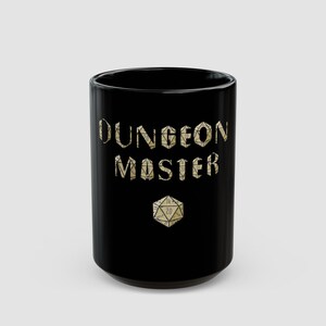 May include: A black ceramic mug featuring the text DUNGEON MASTER printed in a beige, textured font that resembles the surface of polyhedral dice. Below the text, a single twenty-sided die graphic is displayed in the same beige color scheme.