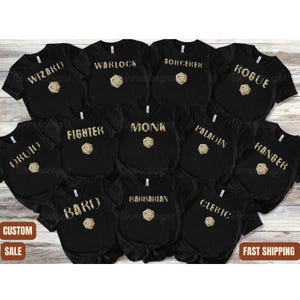 May include: A set of 12 black t-shirts with gold glitter designs. Each shirt features a different Dungeons and Dragons character class, including Wizard, Warlock, Sorcerer, Rogue, Druid, Fighter, Monk, Paladin, Ranger, Bard, Barbarian, and Cleric. The shirts are arranged in a grid pattern with the text "CUSTOM SALE" and "FAST SHIPPING" at the bottom.