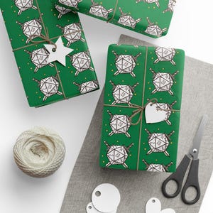 May include: Green gift wrap featuring a repeating pattern of white d20 dice with swords. The wrapped gifts are tied with twine and have white tags. A ball of twine, scissors, and blank tags are also visible.