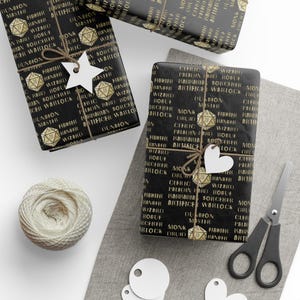 May include: Black wrapping paper with gold text featuring fantasy role-playing game class names and dice. The wrapped gifts are tied with twine and adorned with white star and heart-shaped tags. A ball of twine and scissors are also visible.