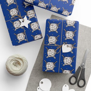 May include: Gift-wrapped presents with blue wrapping paper featuring a white and black d20 dice pattern, crossed swords, and gold stars. The gifts are tied with twine and have blank tags. A ball of twine, scissors, and blank tags are also visible.
