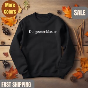 May include: Black sweatshirt with the text "Dungeon Master" in white. The text is centered on the chest. The sweatshirt is surrounded by fall leaves and pinecones. The words "More Colors" and "Sale" are also visible.