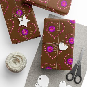 May include: Brown wrapping paper with a repeating pattern of pink hearts and purple twenty-sided dice. The wrapped gifts are tied with twine and adorned with white heart-shaped tags. A spool of twine and scissors are also visible.