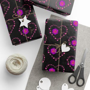 May include: Wrapped gifts with black paper featuring a pattern of pink hearts and purple polyhedral dice. The gifts are tied with twine and adorned with white tags. A ball of twine and scissors are also visible.