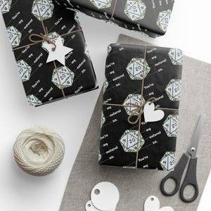 May include: Gift-wrapped presents with black paper featuring white dice and text that reads "You're my natural 20." Tied with twine and adorned with white tags. A ball of twine, scissors, and blank tags are also visible.