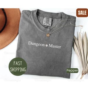 May include: A charcoal gray short-sleeve t-shirt with a crew neckline featuring the white text Dungeon Master with a small dice icon in the center. The shirt has a Comfort Colors label at the inner neck and is displayed with a gold chain necklace.
