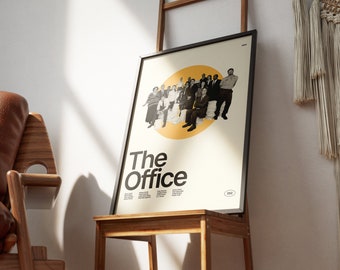 May include: A black framed poster with a white background and a yellow circle in the center. The circle contains a group of people in black and white. The text "The Office" is in black letters below the circle. The poster is on a wooden easel.