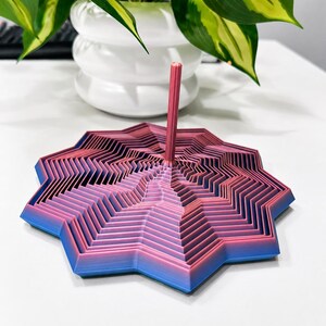 May include: A layered, geometric art piece in shades of pink and blue, with a central pink rod. The design has a star-like shape with a gradient color effect. The piece is likely a decorative item or a unique desk accessory.