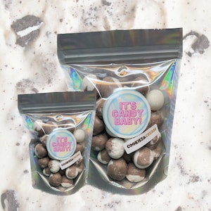 May include: Two clear plastic bags filled with chocolate and white candy balls. The bags have a holographic iridescent finish. The larger bag has a sticker that says "It's Candy Baby!" and "Cookies & Cream". The smaller bag has a sticker that says "It's Candy Baby!" and "Cookies & Cream".