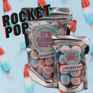 May include: Two clear bags of Rocket Pop candy, each filled with blue and pink candies. The bags have circular labels that read "It's Candy Baby!" and "Rocket Pop." The background features popsicle treats.