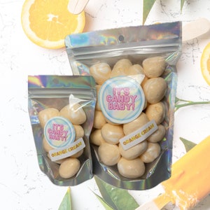 May include: Two bags of orange cream candy. The bags are clear and have a holographic iridescent finish. The candy is white with a yellow swirl and is shaped like a ball. The bags have a sticker that says "It's Candy Baby!" and "Orange Cream".