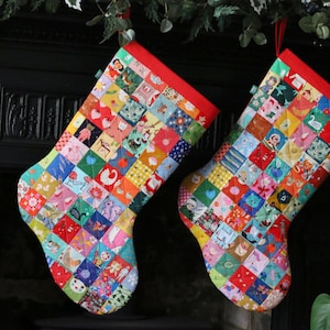 Puede incluir: Dos calcetines navideños con un diseño de patchwork. Cada calcetín está hecho de pequeños cuadrados coloridos con varios patrones e imágenes. La parte superior de cada calcetín es roja y cuelgan de cintas rojas.