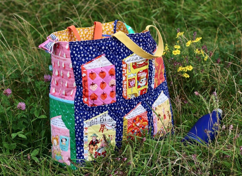 Op de afbeelding: Een kleurrijke patchwork tote bag met diverse stofpatronen, waaronder aardbeien, sterren en vintage-stijl illustraties. De tas heeft meerdere zakken en een gele band, tegen een achtergrond van groen gras en wilde bloemen.