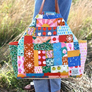 Op de afbeelding: Een kleurrijke patchwork tote bag met diverse patronen, waaronder eekhoorns, ijsjes en bloemmotieven. De tas heeft bruine leren handvatten en een rechthoekige vorm. De tekst "SQUARE STORIES" is zichtbaar aan de zijkant.