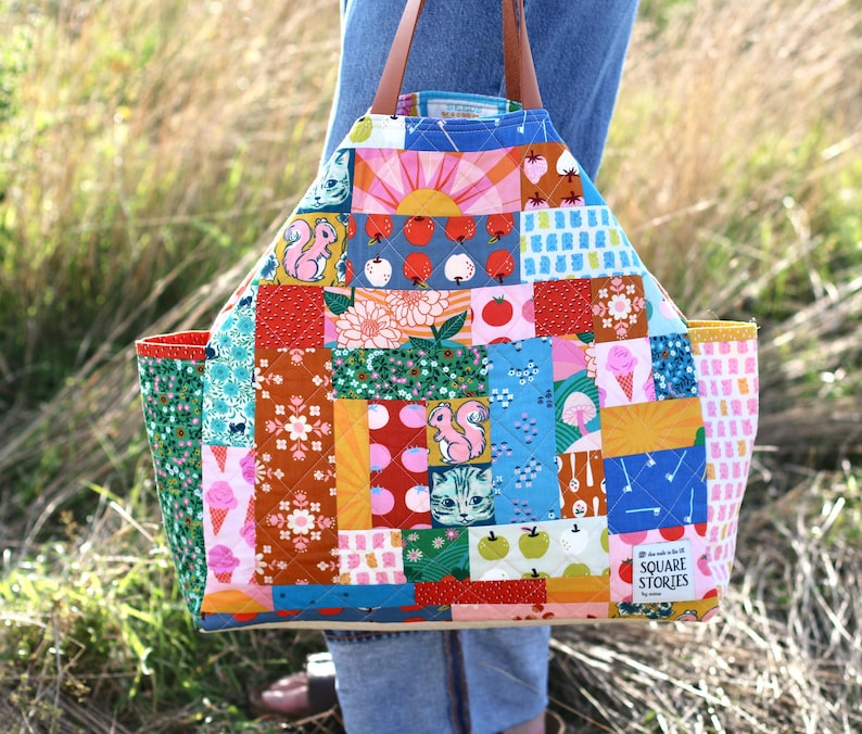 Op de afbeelding: Een kleurrijke patchwork tote bag met diverse patronen, waaronder eekhoorns, ijsjes en bloemmotieven. De tas heeft bruine leren handvatten en een rechthoekige vorm. De tekst "SQUARE STORIES" is zichtbaar aan de zijkant.