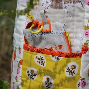 Op de afbeelding: Een gewatteerd stoffen item met een gele zak met een bloemenpatroon. Het item heeft een witte achtergrond met aardbeien- en huisillustraties. Het bevat een zak met ritssluiting en oranje en gele accenten.