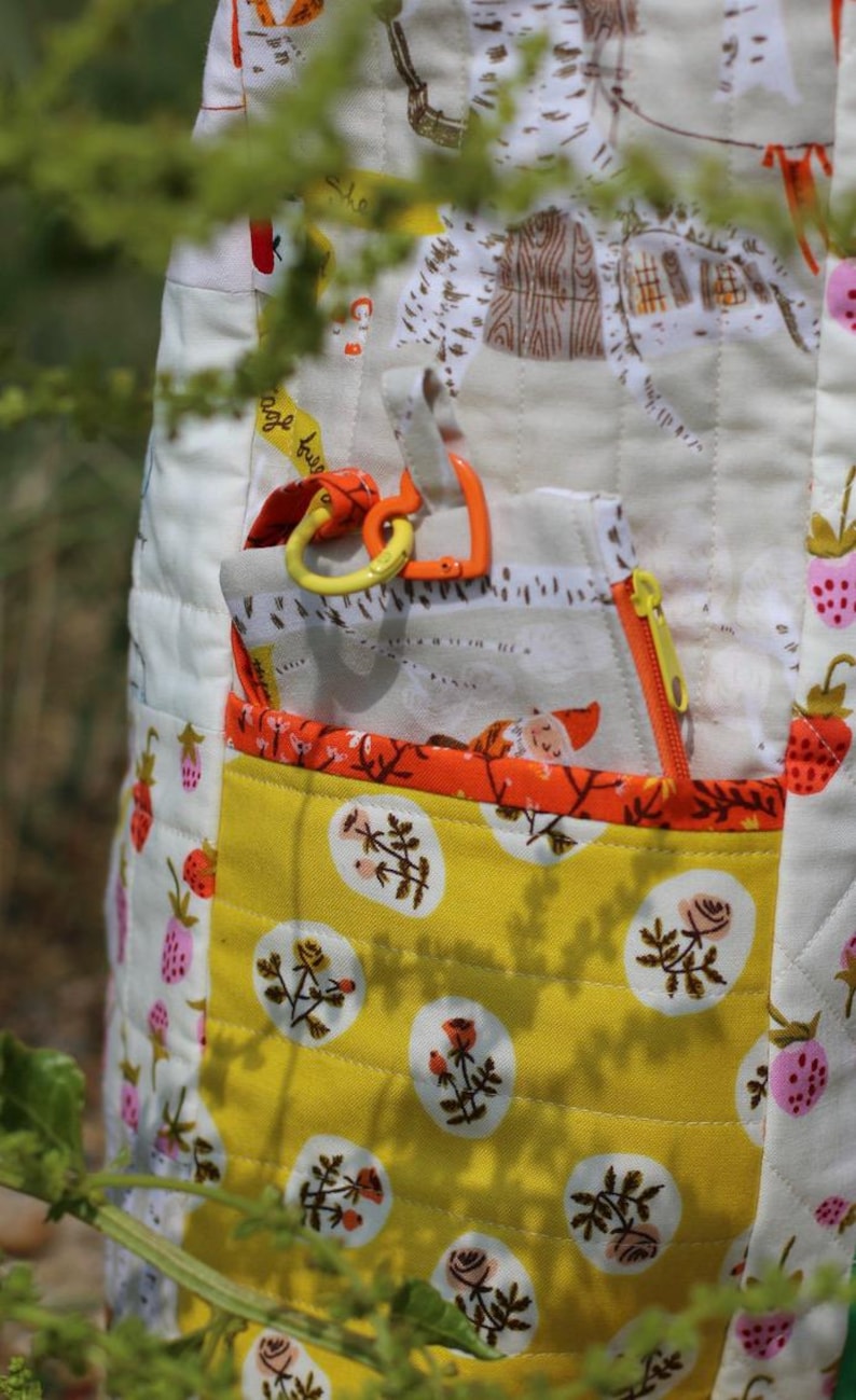 Op de afbeelding: Een gewatteerd stoffen item met een gele zak met een bloemenpatroon. Het item heeft een witte achtergrond met aardbeien- en huisillustraties. Het bevat een zak met ritssluiting en oranje en gele accenten.
