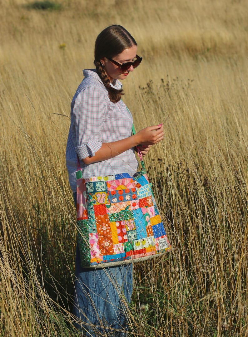 Op de afbeelding: Een kleurrijke patchwork tote bag met diverse stofpatronen, waaronder bloemen- en geometrische ontwerpen. De tas wordt gedragen door een persoon die een geruit overhemd en een spijkerbroek draagt. De tas heeft groene bandjes.