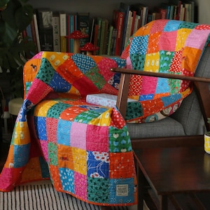 May include: A vibrant patchwork quilt in shades of red, orange, blue, and green, draped over a grey armchair. The quilt is made up of various patterned squares. A small side table and a potted plant are also visible.