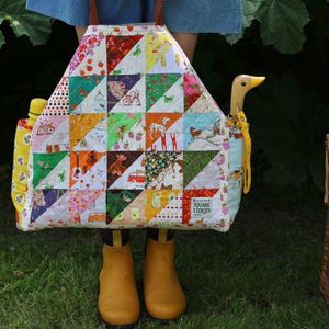 Op de afbeelding: Een kleurrijke patchwork-draagtas met verschillende patronen, waaronder aardbeien, dieren en geometrische vormen. De tas heeft een houten eendvormige handgreep en wordt vastgehouden door een persoon met gele laarzen. Een waterfles is zichtbaar.