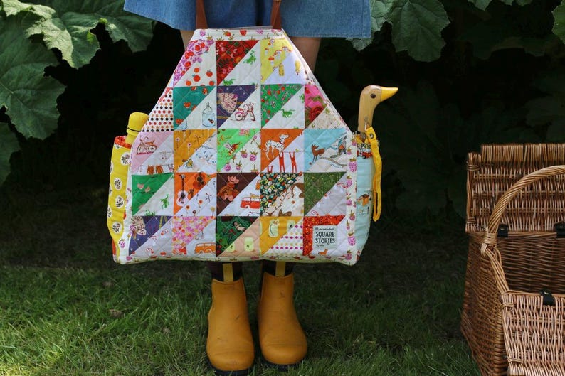 Op de afbeelding: Een kleurrijke patchwork-draagtas met verschillende patronen, waaronder aardbeien, dieren en geometrische vormen. De tas heeft een houten eendvormige handgreep en wordt vastgehouden door een persoon met gele laarzen. Een waterfles is zichtbaar.