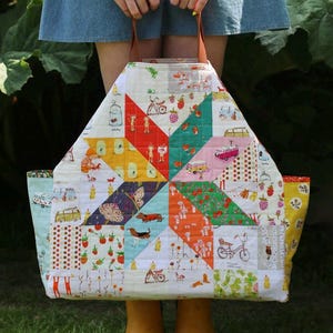 Op de afbeelding: Een grote, gewatteerde draagtas met een patchwork-ontwerp. De tas heeft een verscheidenheid aan kleurrijke patronen, waaronder aardbeien, fietsen en vlinders. Het heeft bruine leren handvatten.