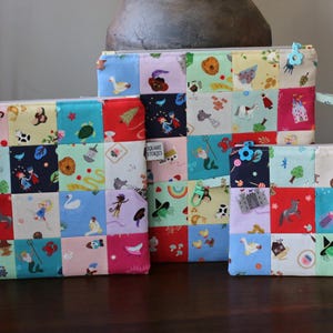 May include: Three rectangular zipped pouches in varying sizes, each featuring a patchwork design of colourful squares with diverse cartoon illustrations. Each pouch includes a small fabric tag labelled Square Stories and a plastic zip pull.