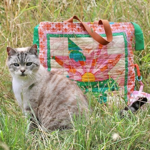Op de afbeelding: Een grijze en witte kat zit in hoog gras, met een kleurrijke gewatteerde draagtas erachter. De tas heeft een groot bloemmotief in roze, groen en oranje. De tas heeft bruine leren handvatten.