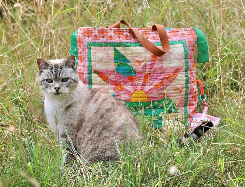 Op de afbeelding: Een grijze en witte kat zit in hoog gras, met een kleurrijke gewatteerde draagtas erachter. De tas heeft een groot bloemmotief in roze, groen en oranje. De tas heeft bruine leren handvatten.
