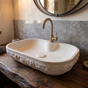 May include: An oval-shaped stone vessel sink with a rough, natural rock-textured exterior and a smooth interior basin. The sink features a central drain and is fitted with a tall, curved, brushed brass faucet with a single lever handle.