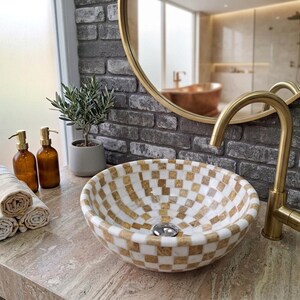 May include: A round vessel sink featuring a white and gold checkered pattern, paired with a tall brushed gold faucet. The sink sits on a stone countertop next to two amber glass soap dispensers, a small potted plant, and three rolled towels.
