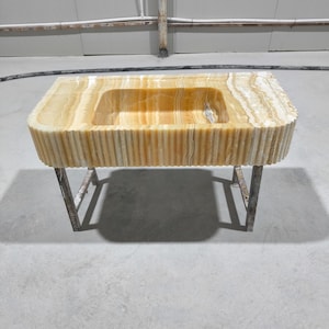 Peut inclure: Un évier rectangulaire en onyx avec un bassin intégré. L'évier a un bord cannelé et est soutenu par un cadre métallique. La pierre présente des tons chauds de jaune, de brun et de blanc.