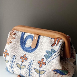 May include: A beige textured fabric pouch featuring colourful embroidered floral and bird motifs in blue, orange, and green. The pouch has a tan leather frame closure with a small tab and a silver metal snap, accompanied by two wooden knitting needles.