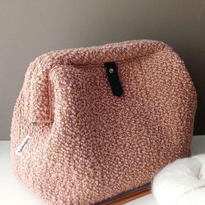 May include: A dusty pink textured pouch with a fuzzy, boucle-like surface. It features a black vertical tab with a silver metal snap closure at the centre. A small white tag with the text Agadesign is attached to the side. Two wooden knitting needles and a ball of white yarn are placed in front.