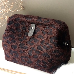 May include: A textured pouch featuring a dark brown and black leopard print pattern. The bag has a rectangular shape with a fold-over top secured by a small black leather-like tab with a silver-toned metal snap button closure.