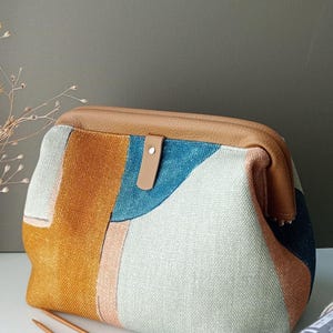 May include: A structured pouch with a tan leather frame and a central tab closure. The body features a textured fabric with an abstract pattern in shades of mustard yellow, teal blue, light grey, and peach. Two wooden knitting needles are visible.