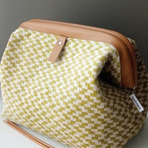 May include: A structured pouch with a tan leather frame and a small tab closure. The body features a woven olive green and cream houndstooth pattern. A white fabric tag with the text Agadesignn is attached to the side. Two wooden knitting needles lie in front.