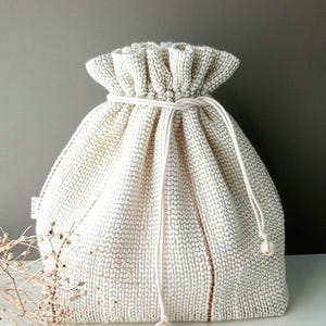 May include: A textured, off-white drawstring bag with a gathered top. The bag features a white drawstring closure with wooden bead accents. Vertical brown stripes add detail to the bag's design. The bag is made of a woven fabric.
