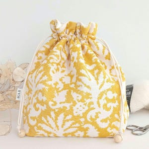 May include: A yellow drawstring bag with a white floral pattern. The bag is made of fabric and has a drawstring closure. The bag is sitting on a white surface.