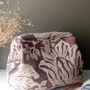 May include: A structured fabric pouch featuring a dark plum and cream botanical print. The bag has a rectangular frame opening with a central silver-toned metal snap closure and a small side tag labelled Agadesigns, positioned near knitting needles.