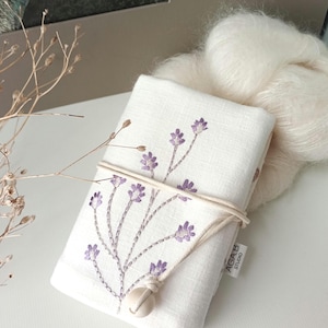 May include: A white linen pouch with embroidered purple floral sprigs, tied with a natural cord and a wooden bead closure. A fluffy, off-white yarn ball rests beside it. The pouch has a small tag that reads "AGAB STUDIO."