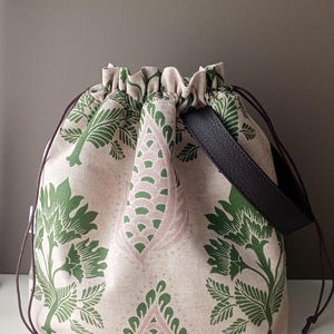 May include: A drawstring pouch bag featuring a beige fabric base with a repeating green and pink botanical print. The bag includes a dark brown leather handle and thin brown drawstring cords. Two wooden knitting needles lie in front of the bag.