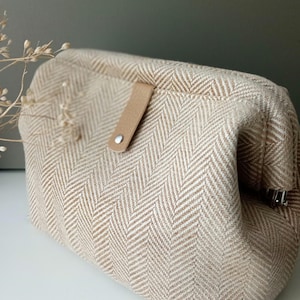 May include: A beige and white herringbone pattern clutch bag with a leather strap and silver-tone metal hardware. The bag is rectangular with a rounded top and a flap closure. The neutral colors and classic pattern give it a timeless look.