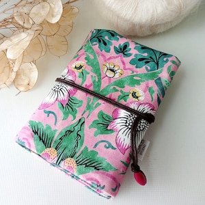 May include: A pink floral case with green leaves and white flowers, fastened with a brown cord and a red bead. The case has a repeating pattern. A ball of white yarn is in the background.