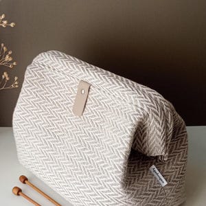 May include: A structured fabric pouch featuring a beige and white herringbone pattern. The bag has a rectangular shape with a fold-over top secured by a light tan leather tab with a silver metal rivet. A small white label reads Agadesign.