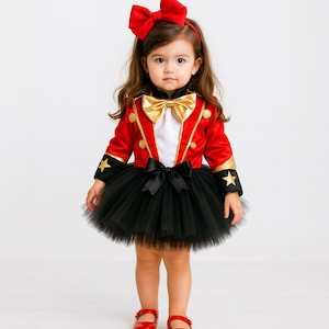 May include: A child's circus-themed costume. The outfit includes a red jacket with gold trim and star accents, a black tutu skirt, and a red bow headband. The ensemble is completed with red shoes and a gold bow tie.