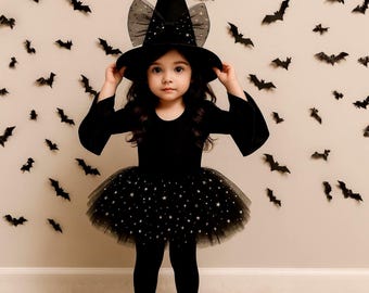 May include: A child's Halloween witch costume. The outfit includes a black velvet dress with bell sleeves, a star-studded tulle skirt, and a pointed witch hat with a bow. The background features bat decorations.